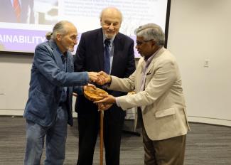 Two men shaking hands as an award is presented. Another man stands behind. 