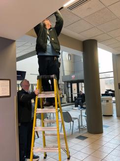Two Facilities team members use a ladder to verify damper orientation on a valve in the ceiling