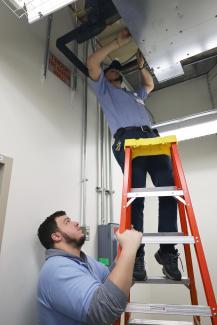Two facilities maintenance technicians, one on a ladder and one holding the ladder, conducting fan coil preventative maintenance