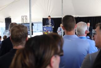 Man speaking at podium in crowd under a tent outside