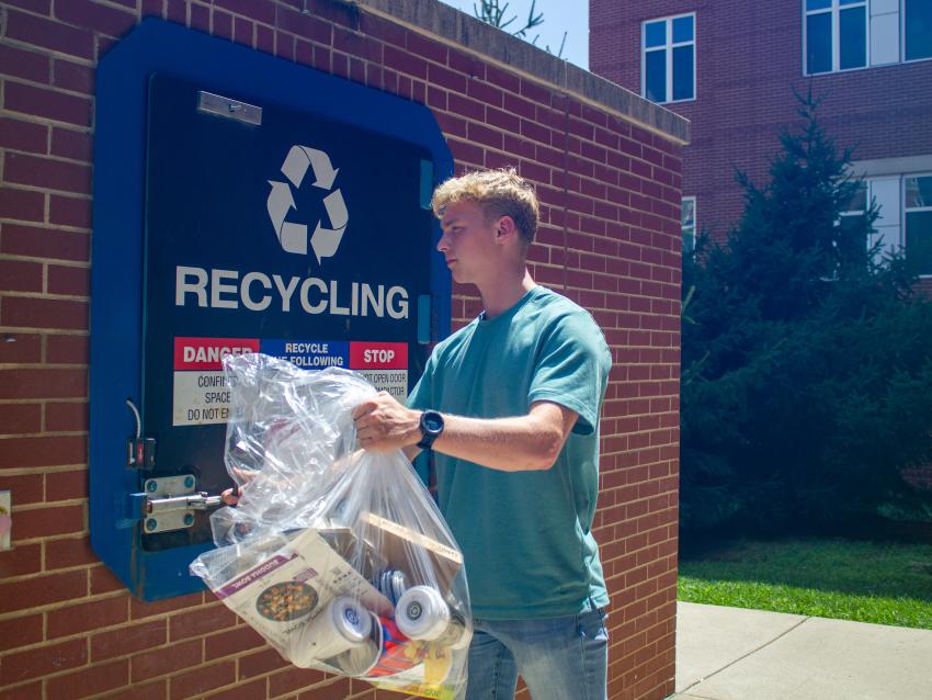 A student takes a clear bag of recyclables to the recycling compactor